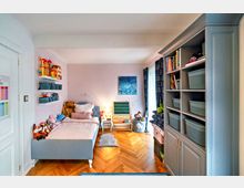 Kinderzimmer mit pastellvioletter Wand, einem Einzelbett mit Stofftieren, Wandregalen mit Spielzeug und Aufbewahrungskörben. Rechts steht ein grauer Schrank mit Büchern und Körben, daneben eine Spieltafel und ein großes Fenster mit gemusterten Vorhängen.