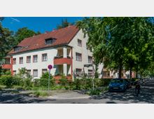 Mehrfamilienhaus mit heller Fassade, roten Fensterrahmen, Balkonen und einem roten Satteldach, umgeben von Bäumen und Grünflächen. Im Vordergrund verläuft eine Straße mit parkenden Autos, einem Verkehrsschild und einem Fahrradfahrer.