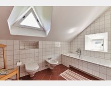 Badezimmer mit Dachschräge, weiß gefliesten Wänden und roten Bodenfliesen. Ausstattung: Badewanne, WC, Bidet, dreieckiges Dachfenster sowie ein kleines rechteckiges Fenster; daneben ein Holzstuhl und ein gestreifter Teppich.