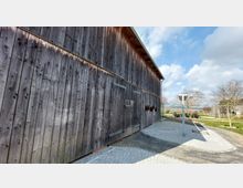 Holzfassade eines großen Gebäudes mit einfacher Bauweise, flankiert von einem gepflasterten Weg und einer Grünanlage mit Bäumen und Sitzbänken im Hintergrund. Der Himmel ist bewölkt mit sonnigen Abschnitten.