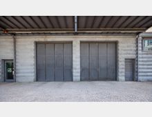 Außenansicht einer grauen Industrietorfassade mit zwei großen Metalltoren und einem kleinen Fenster sowie einer Tür rechts. Der Boden ist mit Kopfsteinpflaster ausgelegt, und ein Überdach aus Metall erstreckt sich über den Eingangsbereich.