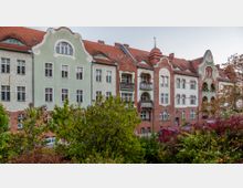 Reihe von mehrstöckigen Wohngebäuden im Jugendstil mit geschwungenen Giebeln, roten Ziegelfassaden und pastellfarbenen Elementen; im Vordergrund Bäume und Sträucher.