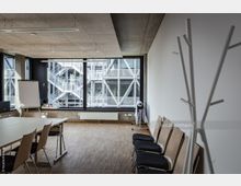 Moderner Seminarraum mit Holzboden, ausgestattet mit einem rechteckigen Tisch, Stühlen, einem Flipchart und einem schlichten Kleiderständer. Große Fenster bieten Ausblick auf ein gegenüberliegendes Gebäude mit Metall- und Glaskonstruktionen sowie Treppen.