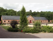 Backsteinbau mit schwarzem Schieferdach, umgeben von einem eingezäunten Sandplatz und Begrünung. Im Hintergrund ist eine dichte Baumreihe zu sehen, die den ländlichen Charakter unterstreicht.