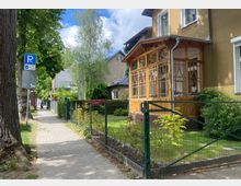 Gehweg entlang einer Wohnstraße mit Einfamilienhäusern, an deren Vorgärten Drahtzäune verlaufen. Rechts im Bild steht ein Haus mit einer verglasten Veranda aus Holz, daneben sind Gehölze und Pflanzen sichtbar.