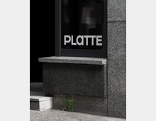 Nahaufnahme einer modernen Gebäudefassade aus grauem Beton mit einem schwarzen Fenster, auf dem der Schriftzug "PLATTE" in weißen Großbuchstaben zu sehen ist; darunter wachsen zwei kleine grüne Pflanzen aus einem Spalt im Boden.