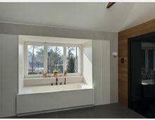 Modernes Badezimmer mit einer großen, weißen Badewanne vor einem breiten Fenster, das Tageslicht hereinlässt. Die Wand rechts ist mit Holzvertäfelung verkleidet, während der Boden aus dunklen Fliesen besteht.