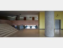 Innenraum eines modernen Gebäudes mit breiter Treppe, einem gelben und einem rosafarbenen Wandbereich. Links führt ein Geländer an der Treppe entlang, in der Mitte ist eine große Betonsäule, rechts eine Wanduhr und ein Aufzug; der Bereich ist als "Unteres Foyer Konferenzräume" ausgeschildert.