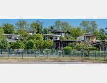 Mehrere kleine Gartenhäuser auf einem terrassenförmigen Hang, umgeben von üppigem Grün, Bäumen und Hecken. Im Vordergrund ein Drahtzaun und ein gepflasterter Weg, dahinter eine Steinmauer, die die Grundstücke abgrenzt.