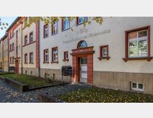 Fassade eines mehrstöckigen Wohnhauses mit beiger und roter Farbgestaltung, mehreren weißen Fenstern und einer Tür mit der Hausnummer 100. Vor dem Gebäude befindet sich ein kleiner Grünstreifen mit einem gepflasterten Weg und einem Briefkastenständer.