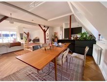 Ein modernes Wohnzimmer im offenen Stil mit sichtbaren Holzbalken, Parkettboden und verschiedenen Teppichen. Im Vordergrund steht ein Esstisch mit Stühlen, im Hintergrund sind eine schwarze Küchenzeile, ein Sofa und große Fenster mit Blick nach draußen sichtbar.