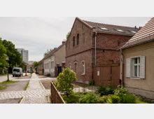 Gepflasterte Straße in einer Wohngegend mit historischen Backstein- und Putzgebäuden. Im Vordergrund ein kleiner bepflanzter Vorgarten, im Hintergrund moderne Hochhäuser und Bäume.