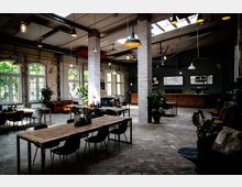 Ein großzügiges Café oder Restaurant in einem industriellen Stil mit freiliegenden Backsteinwänden, großen Fenstern und sichtbaren Deckenbalken. Das Interieur umfasst Holztische, schlichte Stühle, hängende Lampen und grüne Pflanzen, die den Raum einladend wirken lassen.
