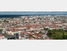Panoramaansicht einer Stadt mit dicht bebauten Wohngebieten, überwiegend mit roten Ziegeldächern. Im Vordergrund ist eine Hauptstraße zu sehen, dahinter erstrecken sich urbane Gebäude und einzelne grüne Flächen, während am Horizont Industriebauten und Windräder sichtbar sind.