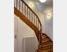 Holztreppe mit sichtbaren Gebrauchsspuren in einem Innenraum mit weiß gestrichenen Wänden, einem Heizkörper unterhalb der Treppe und zwei Wandlampen zur Beleuchtung.