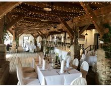 Rustikal eingerichteter Saal mit Holzbalkendecke und dekorativen Lichterketten. Tische sind mit weißen Tischdecken, gefalteten Servietten und floralen Arrangements gedeckt, umgeben von gemauerten Säulen und Holzböden.