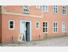 Außenansicht eines historischen Gebäudes mit rosafarbener Fassade, weißen Fenstern und einer geöffneten Doppeltür mit verzierten Türrahmen. Vor der Tür steht ein Aufsteller mit Informationen, der Eingangsbereich liegt an einem gepflasterten Boden.