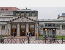 Eingang zum U-Bahnhof Wittenbergplatz in Berlin mit einer historischen Fassade aus grauem Stein, flankiert von zwei verzierten Türen. Im Vordergrund ein Zaun, Fahrräder und eine moderne Aufzugsanlage mit Glasdach, im Hintergrund ein größeres Gebäude mit rotem Dach.