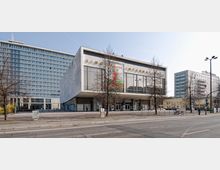 Außenansicht des Kinos International in Berlin, einem rechteckigen Gebäude mit großflächigen Glasfenstern und einem leicht vorspringenden Dach. Im Hintergrund befinden sich moderne Wohn- und Bürogebäude, während einige kahle Bäume und eine Straße den Vordergrund prägen.