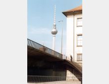 Straßenansicht mit dem Berliner Fernsehturm im Hintergrund, einer Brücke im Vordergrund und einem Teil eines Gebäudes mit roten Dachziegeln und mehreren Fenstern rechts. Eine Person geht eine Treppe hoch, die an die Brücke grenzt.