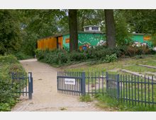 Ein Sandweg führt durch ein kleines Metalltor mit einem Schild „Hunde verboten“ zu einem einstöckigen Gebäude mit bunter Wandmalerei, umgeben von Bäumen und Sträuchern in einer parkähnlichen Umgebung.