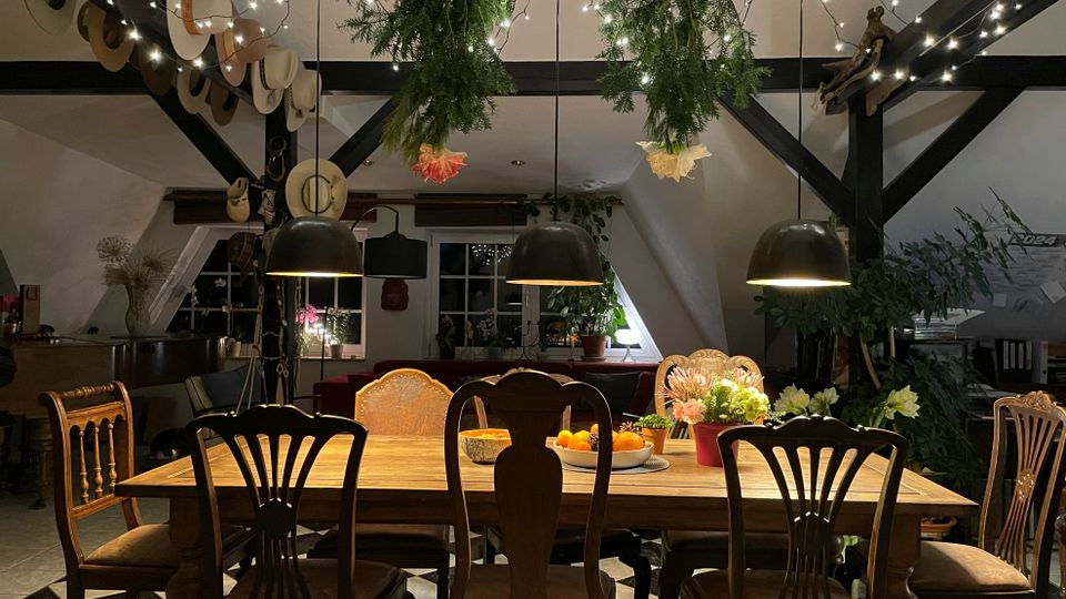 Gemütlicher Essbereich in einem Dachgeschoss mit sichtbaren schwarzen Holzbalken, großem Holztisch und verschiedenen Stühlen auf einem schwarz-weiß karierten Boden. Über dem Tisch hängen drei schwarze Pendelleuchten, Lichterketten und aufgehängtes Grün; im Hintergrund sind Fenster, Pflanzen und Dekoration zu sehen.