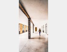 Großer, heller Industrie-Loft-Innenraum mit sichtbaren Stahlträgern und Backsteinwänden entlang der linken Seite. Hohe Fensterreihen und Türen auf beiden Seiten sowie ein glatter Betonboden prägen den offenen Raum.