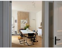 Blick durch einen Türrahmen in ein helles Esszimmer mit großem rechteckigem Tisch, schwarzen Stühlen und hängender Pendelleuchte. Im Hintergrund stehen eine freigelegte Ziegelwand mit Poster, ein Sideboard sowie eine Vase mit Blumen auf dem Tisch auf hellem Holzfußboden.