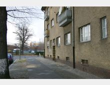 Straßenansicht mit einem mehrstöckigen Wohngebäude aus verputztem Beton, das kleine Balkone und rechteckige Fenster hat. Im Vordergrund Gehweg mit Pflastersteinen, parkende Autos und ein Baum; im Hintergrund sind vereinzelte Bäume, Verkehrsflächen und moderne Gebäude zu sehen.