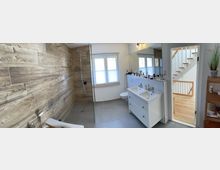 Modernes Badezimmer mit großer begehbarer Dusche hinter Glas, Holzoptik-Wandfliesen und grauen Bodenfliesen. Rechts ein weißer Doppelwaschtisch mit Spiegel, daneben ein Fenster; durch die offene Tür ist ein heller Flur mit Holztreppe sichtbar.