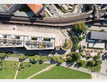 Luftaufnahme eines städtischen Gebiets mit einer Mischung aus Grünflächen, modernen Gebäuden mit begrünten Dächern und einem Parkplatz. Oben im Bild verlaufen Bahngleise neben mehrstöckigen Ziegelgebäuden, während unten ein Park mit Wegen und Bäumen sichtbar ist.