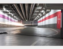 Ein beleuchteter Tiefgaragenbereich mit geschwungener Fahrbahn, Lüftungsrohren an der Decke und Wandstreifen in Weiß, Rot und Grau. Ein heller Lichtstreifen, vermutlich von einem fahrenden Fahrzeug, zieht sich entlang der Straße.