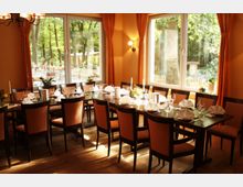 Innenraum eines Restaurants mit gedecktem Tisch für mehrere Personen. Große Fenster mit orangefarbenen Vorhängen bieten Blick auf eine grüne Außenanlage, während das warme Licht eine gemütliche Atmosphäre erzeugt.