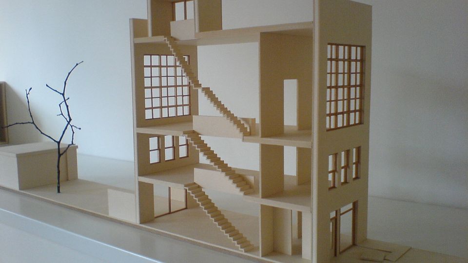 A minimalist architectural model of a multi-story building with open walls, featuring a series of staircases connecting the floors, large grid-style windows on each level, and a plain base with a small branch-like structure to the side.
