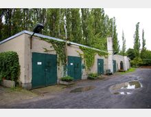 Eine Reihe von Beton-Garagen mit grünen Metalltoren, teils bewachsen mit Efeu. Im Vordergrund ist ein asphaltierter Hof mit Pfützen, im Hintergrund stehen hohe Bäume.