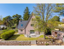 Freistehendes Einfamilienhaus mit steilem Satteldach und hellrosa Fassade, davor ein gepflegter Garten mit Terrasse, Rasenfläche, niedriger Steinmauer und Treppenstufen. Umgeben von hohen Bäumen und Hecken in einer ruhigen Wohngegend bei blauem Himmel.