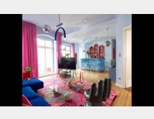 Wohnzimmer in einem modernen, farbenfrohen Stil mit hellblauen Wänden, rosa Vorhängen und Holzboden. Im Raum befinden sich ein blauer Sofa, ein Fernseher, ein Glas-Couchtisch auf einem bunten Teppich und eine Bar mit glänzender, blauer Verkleidung und bogenförmigen Regalen im Hintergrund.