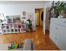 Wohnzimmer mit Parkettboden, einer grauen Couch im Vordergrund und einem Spielzeug auf einem kleinen Teppich. An der Wand stehen weiße Bücherregale mit Büchern und Dekoration, darüber hängen Bilder. Rechts befinden sich eine weiße Kommode und Zimmerpflanzen, die Tür führt zu einem Flur.
