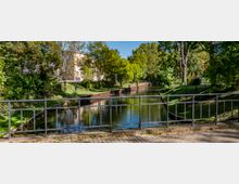 Eine kleine Wasserstraße, gesäumt von Bäumen und einer begrünten Uferpromenade, mit einem Metallgeländer im Vordergrund. Im Hintergrund sind ein mehrstöckiges Gebäude und ein Parkplatz erkennbar.