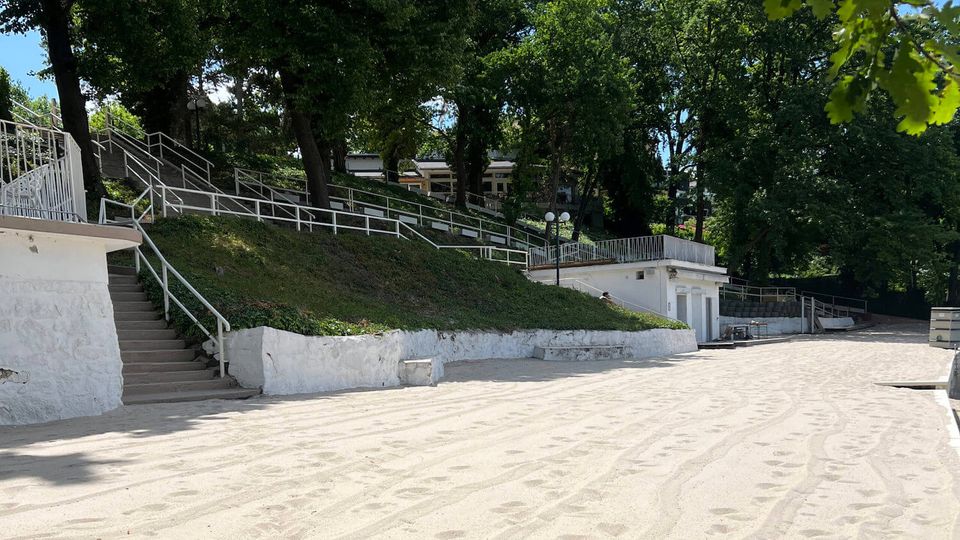 Sandy open area bordered by low white retaining walls and a sloped hillside with stairs and white railings. Dense trees line the upper terrace, with small white utility buildings and lampposts visible along the path.