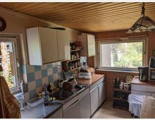 Kleine, rustikale Küche mit Holzdecke und holzvertäfelten Wänden, weißen Hängeschränken und blau-weiß gefliester Rückwand über einer langen Arbeitszeile mit Spüle. Rechts sorgt ein großes Fenster für Tageslicht; auf den Arbeitsflächen stehen Küchengeräte und offene Regale mit Utensilien.