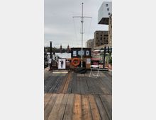 Holzstege eines Bootsanlegers mit einer kleinen Kabine, Rettungsring und Tau im Vordergrund. Im Hintergrund die Oberbaumbrücke, flankiert von modernen Gebäuden und einem bewölkten Himmel.