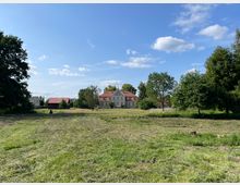 Weite Wiese mit frisch gemähtem Gras, umgeben von Bäumen. Im Hintergrund steht ein großes Gebäude mit heller Fassade und rotem Dach, daneben einige kleinere Häuser und Scheunen mit roten Dächern, unter blauem Himmel mit wenigen Wolken.