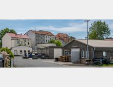 Hofbereich mit mehreren Gebäuden: links Wohnhäuser mit roten Ziegeldächern, rechts eine Lagerhalle mit Wellblechverkleidung. Im Vordergrund Müllcontainer, Holzpaletten und abgestellte Fahrzeuge, im Hintergrund Bäume und blauer Himmel.