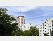 Mehrstöckige Wohngebäude in einer urbanen Umgebung, darunter ein hohes weiß-rotes Hochhaus mit markantem Design. Im Vordergrund befinden sich Bäume, Straßenlaternen und ein gelber Linienbus.