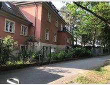 Zweistöckiges Wohngebäude mit rosafarbener Fassade und weißen Sprossenfenstern, umgeben von einem Garten mit Zaun. Im Hintergrund sind hohe Bäume und eine gepflasterte Gehwegfläche zu sehen.