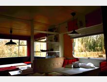 Innenansicht eines Wohnwagens mit gemütlicher Sitzecke, bestehend aus einer gepolsterten Bank und einem weißen Tisch. Große Fenster mit Blick auf eine bewaldete Außenlandschaft lassen Tageslicht in den Raum, der durch Hängelampen und farbige Akzente dekoriert ist.