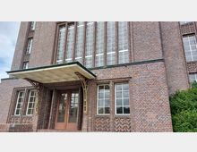 Ansicht eines Backsteingebäudes mit einer überdachten Eingangsfront. Die Architektur zeichnet sich durch große, vertikale Fenster, Ziegelverzierungen und ein grünes Vordach mit metallenen Stützstreben aus.