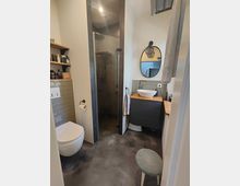 Kleines modernes Badezimmer mit fugenlosem Boden, einer bodengleichen Dusche mit Glastür, einer Wandtoilette, einem ovalen Spiegel über einem Waschbecken auf einem grauen Waschtisch sowie Wandregalen mit Pflegeprodukten. Ein Hocker steht vor dem Waschtisch.