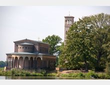 Historische Kirche mit Arkadengang und gestreifter Backsteinfassade, im Vordergrund ein Fluss und Vegetation, im Hintergrund ein freistehender Glockenturm und Bäume.